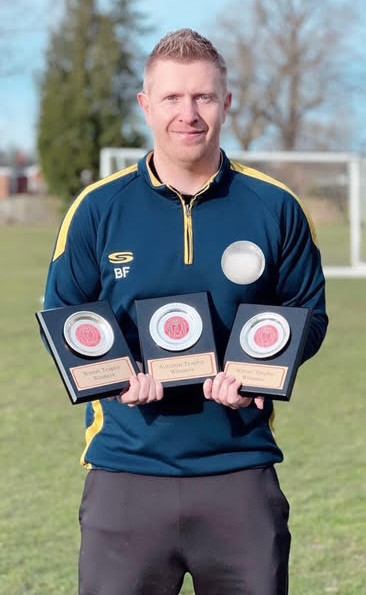 An image of our head coach Brendan holding trophies won by the club